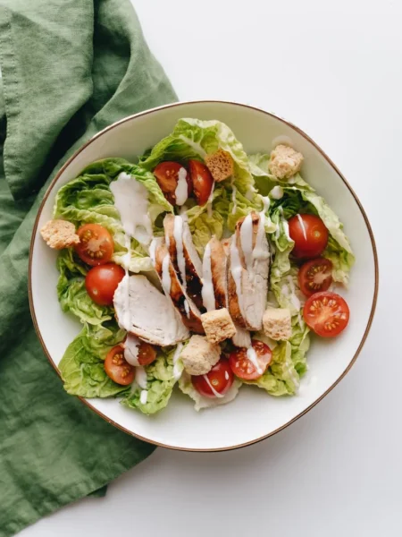 chicken and pesto salad with sliced grilled chicken, basil pesto coated romaine, cherry tomatoes, and toasted pine nuts in a white bowl