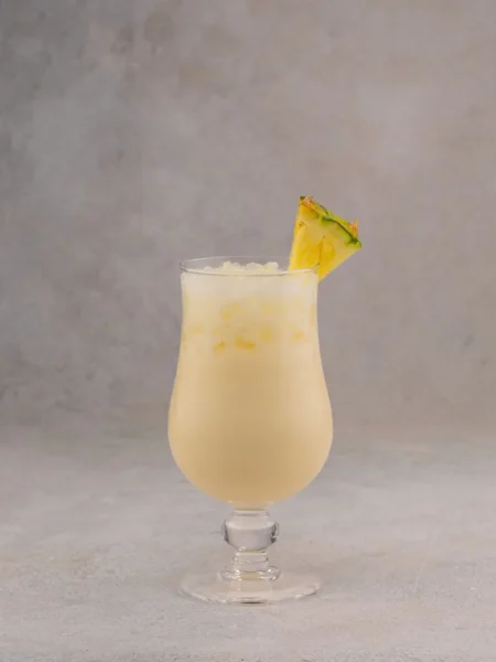 pina colada smoothie in a tall glass with frozen pineapple chunks and coconut foam on top