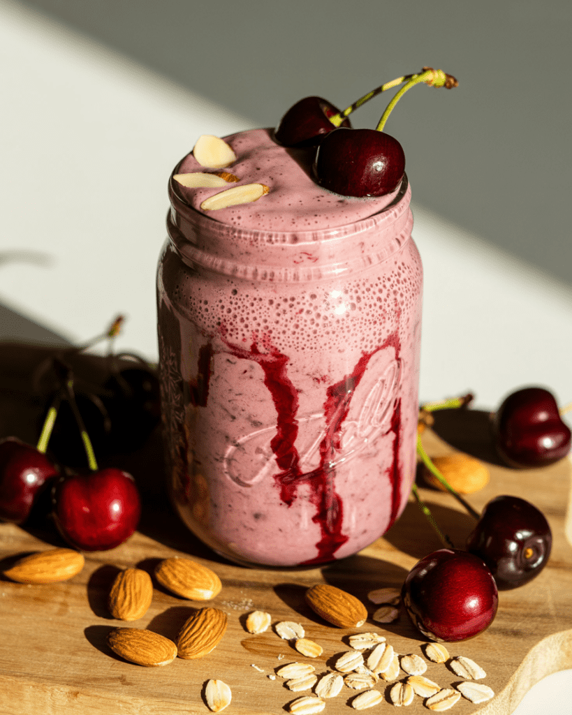 A vibrant food photography shot of a thick, creamy smoothie in a tall glass mason jar, showcasing rich pink and burgundy swirls from fresh cherries blended with creamy almond milk and oats. The smoothie has a luxurious, velvety texture with visible specks of ground almonds and a frothy top, garnished with fresh dark red cherries and sliced almonds on the rim. The glass sits on a rustic wooden cutting board surrounded by scattered whole almonds, fresh cherries with stems, and rolled oats, with soft natural morning light streaming from the left side. The scene conveys wholesome nutrition and artisanal breakfast appeal with warm, inviting tones and clean, minimalist styling.